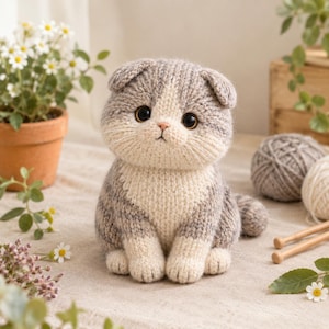 May include: A knitted stuffed toy shaped like a cat with folded ears. The cat has a grey and white striped pattern, large dark brown eyes, and a small pink nose. It is crafted with visible yarn stitches and sits in an upright position.