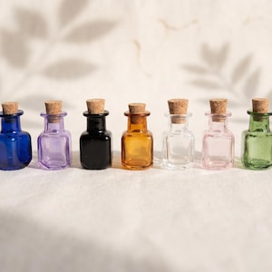 May include: A row of seven small glass bottles with cork stoppers, each featuring a distinct color including blue, purple, black, amber, clear, pink, and green. The bottles have a square base and a narrow neck, arranged in a straight line.