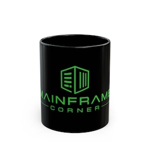 May include: A black ceramic mug featuring a bright green graphic logo on the front. The logo depicts a stylised cube structure above the text MAINFRAME CORNER in a clean, sans-serif font. The mug has a smooth, glossy finish and a cylindrical shape.