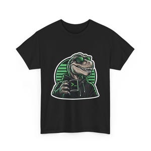 Peut inclure: Un t-shirt noir avec un motif représentant un Tyrannosaure Rex portant des lunettes de soleil et un sweat à capuche noir. Le dinosaure tient une tasse noire avec un symbole de ligne de commande vert. Le fond du motif est un cercle rayé vert.
