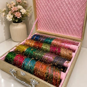 Op de afbeelding: Een rechthoekige goudkleurige opbergkoffer met een roze binnenvoering, gevuld met rijen dunne, veelkleurige glazen armbanden. Elke armband is voorzien van kleine, goudkleurige metalen accenten. De koffer heeft een zilverkleurig metalen handvat.