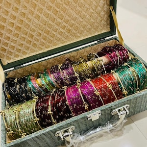 May include: A rectangular storage box with a textured green exterior and a tan interior lining, filled with rows of translucent bangles in various colors including black, green, purple, red, and gold, each featuring small metallic accents.