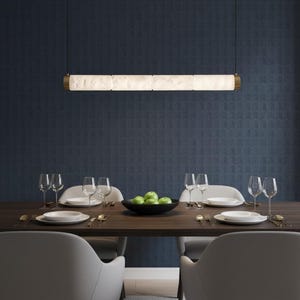 May include: A linear pendant light fixture featuring four cylindrical, textured white translucent segments held by metallic end caps, suspended by thin black wires above a dining table set with plates, glasses, and a bowl of green apples.
