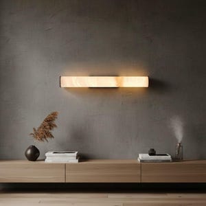 Könnte beinhalten: Eine rechteckige Wandleuchte mit einer warm leuchtenden, durchscheinenden, steinartigen Platte. Die Leuchte strahlt ein sanftes, diffuses Licht an eine dunkelgraue Wand und befindet sich über einem langen, hellen Holzkonsolentisch.