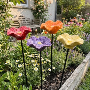 May include: Four ceramic flower garden stakes featuring glossy, ruffled petals in red, orange, purple, and yellow. Each flower is mounted on a thin, black metal rod, designed to be inserted into the ground as a decorative garden ornament.
