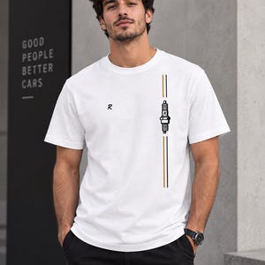 Puede incluir: Una camiseta blanca de manga corta con un diseño gráfico vertical en el lado izquierdo. El diseño incluye una ilustración de una bujía en blanco y negro centrada entre dos finas líneas verticales en dorado y negro, con una letra R impresa.
