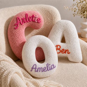 May include: Three plush, textured letter-shaped pillows. A pink letter C features the name Arlette in dark pink embroidery. A white letter O features the name Amelia in purple embroidery. A white letter A features the name Ben in orange embroidery.
