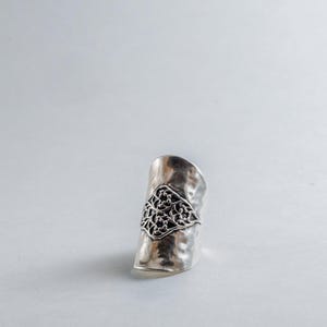 May include: A wide silver ring featuring a hammered, textured surface and a central, raised diamond-shaped panel. The panel displays an intricate, dark, openwork filigree pattern with swirling, vine-like details set against the polished metal.
