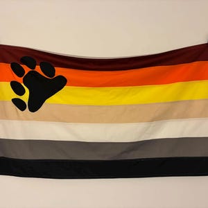 May include: A rectangular flag featuring horizontal stripes in shades of dark brown, orange, yellow, tan, white, gray, and black. A large black bear paw print graphic is positioned on the left side of the flag, overlapping the top four stripes.