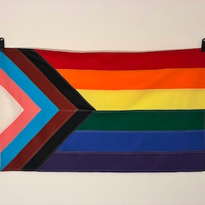 Könnte beinhalten: Eine rechteckige Progress-Pride-Flagge mit horizontalen Streifen in Rot, Orange, Gelb, Grün, Blau und Violett. Auf der linken Seite befindet sich ein Chevron-Muster mit weißen, rosa, hellblauen, braunen und schwarzen Streifen sowie zwei Metallösen.