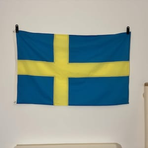 Peut inclure: Un drapeau rectangulaire de la Suède présentant un champ bleu avec une croix nordique jaune. Le drapeau est fixé horizontalement sur un mur à l'aide de deux pinces noires aux coins supérieurs. Une bande blanche avec deux œillets est visible.