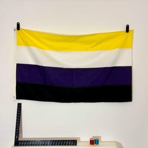 Puede incluir: Una bandera no binaria rectangular con cuatro franjas horizontales en amarillo, blanco, morado y negro. La bandera está sujeta por dos clips negros. Una regla metálica negra en forma de L se sitúa debajo para referencia de escala.