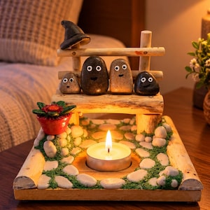 May include: A wooden bench display featuring four smooth stones with googly eyes, a small black witch hat, and a red potted plant. The base is decorated with white pebbles and green moss, centered around a lit white tea light candle.