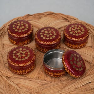 May include: Five round, red glittery containers with metallic gold accents. Four containers have lids featuring a central floral pattern of gold dots and teardrop shapes, with a matching border. One container is open, showing a silver interior.