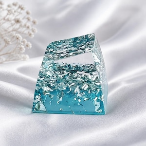 May include: A translucent, trapezoidal mechanical keyboard keycap featuring a gradient of clear to bright blue resin embedded with shimmering silver metallic flakes throughout the structure, creating a reflective and textured appearance.