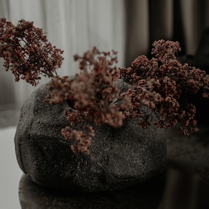 May include: A low, rounded, dark charcoal-colored vase with a rough, textured surface. The vase has a wide opening at the top and contains several sprigs of dried, reddish-brown flowers that extend outward from the center of the vessel.