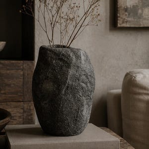 Könnte beinhalten: Eine dunkelgraue, texturierte Steinvase mit unregelmäßiger, organischer Form. Die Vase enthält dünne, getrocknete braune Zweige. Sie steht auf zwei gestapelten Büchern, einem hellgrauen und einem dunkelgrauen mit dem Text wabi inspirations.