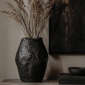 Può includere: Un vaso nero dalla texture organica e irregolare contenente un mazzo di erbe ornamentali essiccate di colore marrone chiaro. Il vaso presenta una superficie ruvida e un corpo ampio e arrotondato che si restringe verso l'apertura superiore.