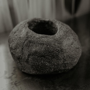 Puede incluir: Un recipiente redondeado de color gris oscuro con una superficie rugosa y texturizada y una abertura circular ancha en la parte superior. El objeto tiene un perfil bajo y achaparrado con un acabado mate, similar a la piedra natural o roca volcánica.