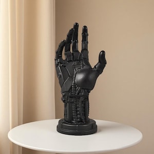 May include: A matte black sculpture of a robotic hand standing upright on a circular base. The hand features intricate mechanical details, including visible joints, segmented fingers, and exposed cable-like textures along the wrist and palm area.