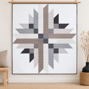 May include: A square wall quilt featuring a central starburst pattern composed of geometric strips in shades of charcoal, slate gray, light gray, and muted brown against a white background, finished with a thin dark gray border.