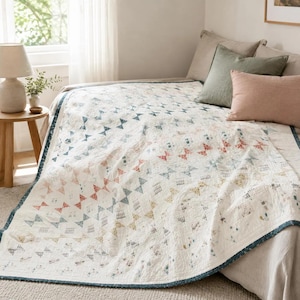 Könnte beinhalten: Eine weiße Steppdecke mit einem sich wiederholenden Muster aus kleinen Dreiecken in gedeckten Blau-, Rot- und Gelbtönen. Die Decke hat einen dunkelblauen Rand und liegt auf einem Bett mit einem salbeigrünen und einem altrosa Kissen.