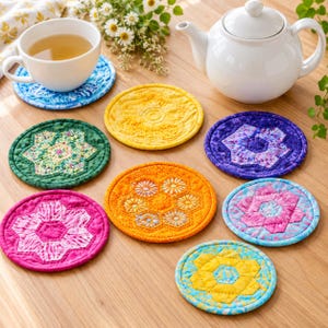 May include: A collection of seven round fabric coasters featuring various quilted hexagonal patterns in vibrant colors including yellow, green, purple, pink, and orange, arranged on a wooden surface alongside a white teapot and teacup.