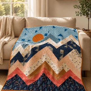 May include: A quilted blanket featuring a mountain landscape design with a bright orange sun, layered peaks in shades of blue, peach, and tan, and a dark navy base patterned with small trees and deer, finished with a solid orange border.