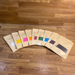 Pode incluir: Nove pequenas bolsas retangulares de papel castanho claro dispostas em leque. Cada bolsa tem uma janela retangular transparente no centro, revelando conteúdos coloridos: branco, amarelo, rosa, laranja, vermelho, verde, azul, roxo e preto.