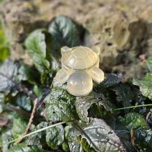 Pode incluir: Uma pequena estatueta de tartaruga translúcida em amarelo pálido com uma carapaça lisa e arredondada e membros definidos. A tartaruga tem um design simples com duas pequenas reentrâncias para os olhos e um padrão segmentado no dorso.