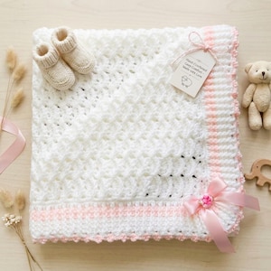 May include: A white crocheted baby blanket with a textured diamond pattern, featuring a light pink border, a pink satin ribbon bow, and a tag reading Hand-Crocheted Cozy Comfort Made with Love. Includes a pair of matching white crocheted booties.