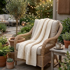 May include: A light beige woven throw blanket with a textured waffle pattern and ribbed edges draped over a natural wicker armchair with a beige cushion. The blanket features a consistent knit design and is positioned centrally on the seat.
