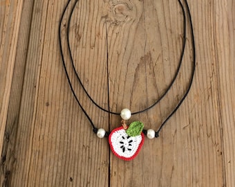 May include: A black cord necklace with a crocheted apple pendant. The apple is red and white with a green leaf and white pearl accents.