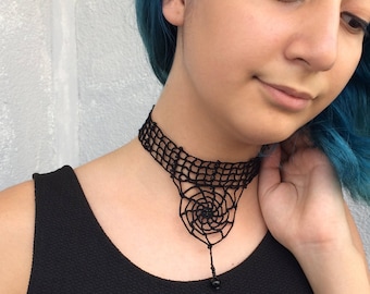 May include: A black crocheted choker necklace with a spiral design and a black bead at the end.