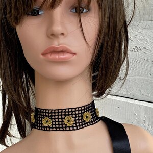 May include: A black crochet choker necklace with gold floral detailing. The choker is worn around the neck and is made of a mesh-like material. The gold flowers are evenly spaced along the front of the choker.