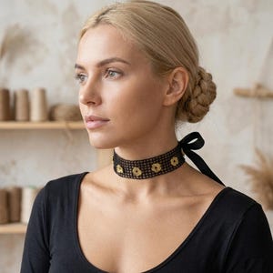 May include: A black choker necklace with a floral design and a black ribbon tie. The necklace is made of a mesh material and features gold-coloured flower accents. The woman in the image is wearing a black top.