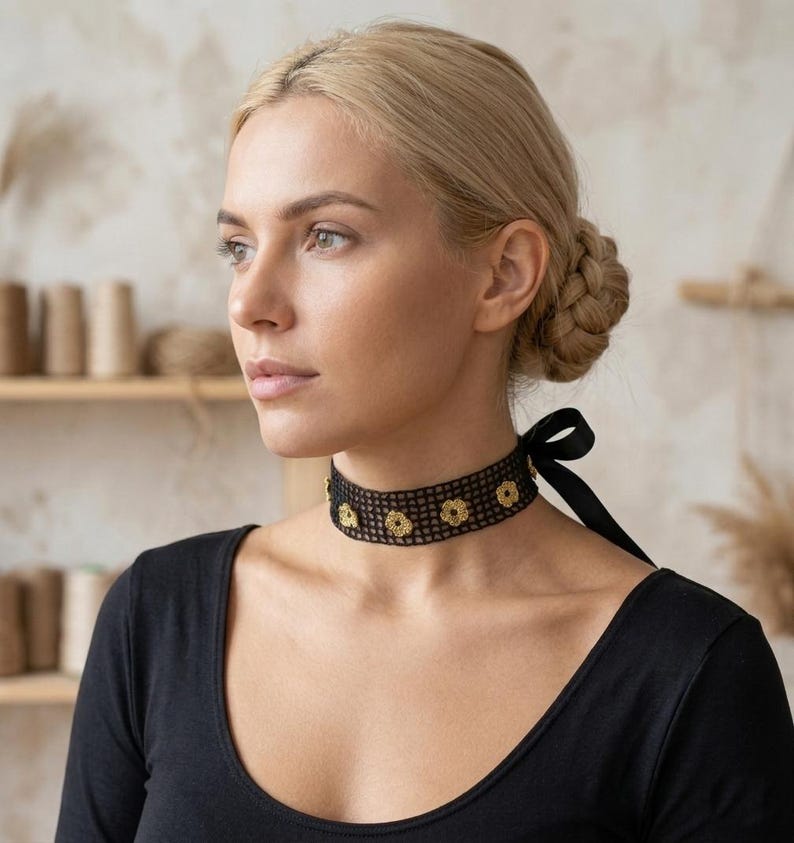 May include: A black choker necklace with a floral design and a black ribbon tie. The necklace is made of a mesh material and features gold-coloured flower accents. The woman in the image is wearing a black top.