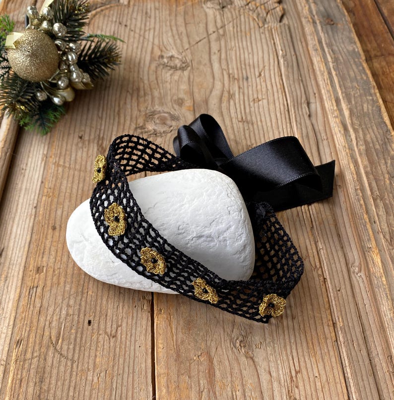 May include: A black crocheted headband with gold flower details, displayed on a white stone. A black satin ribbon is tied in a bow. The headband is presented on a rustic wooden surface, with a Christmas decoration in the background.
