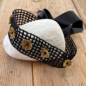 May include: A black crocheted headband with gold-coloured floral accents and a black satin ribbon. The headband is displayed on a white stone against a wooden background. The floral accents are evenly spaced along the headband.