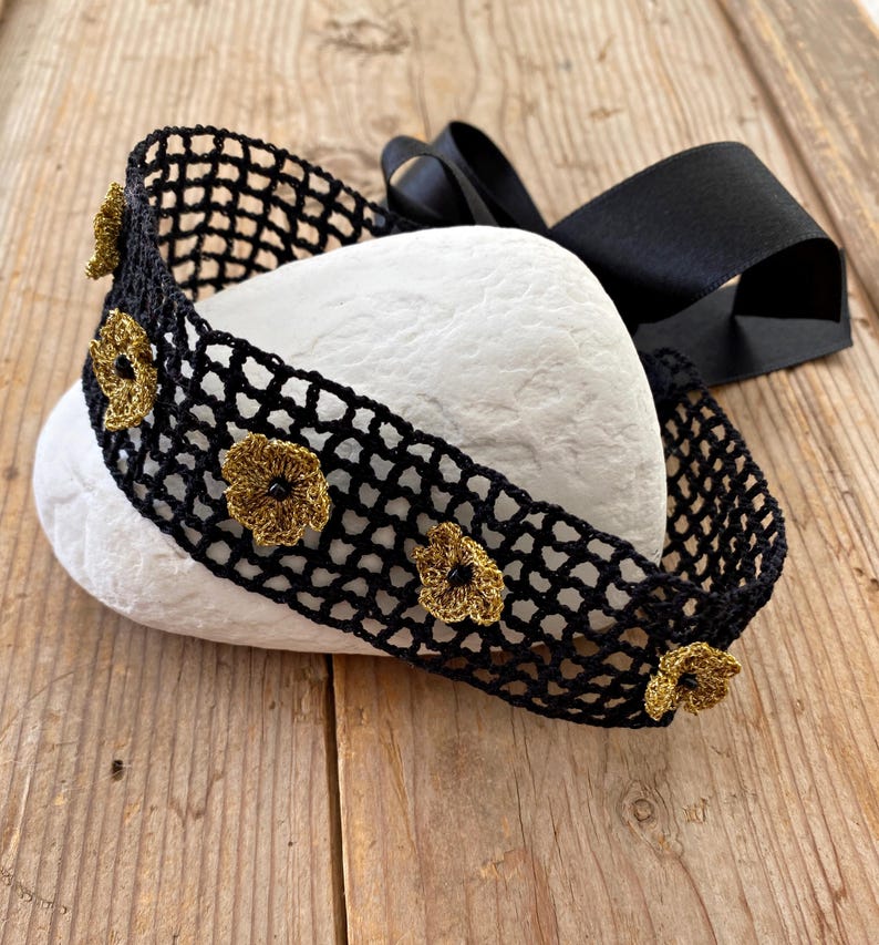 May include: A black crocheted headband with gold-coloured floral accents and a black satin ribbon. The headband is displayed on a white stone against a wooden background. The floral accents are evenly spaced along the headband.