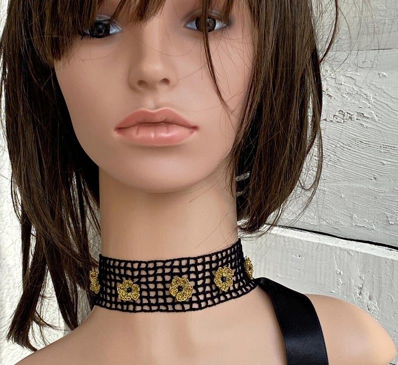 May include: A black crochet choker necklace with gold floral detailing. The choker is worn around the neck and is made of a mesh-like material. The gold flowers are evenly spaced along the front of the choker.