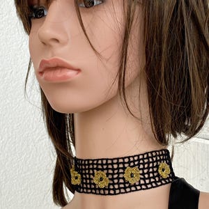 May include: A black crochet choker necklace with gold flower detailing. The necklace is worn around the neck and is made of a woven, open-weave material. The gold flowers are evenly spaced along the front of the choker.