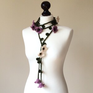 May include: A crocheted flower necklace with purple and white flowers and green leaves. The necklace is displayed on a white mannequin.