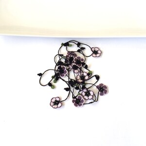 May include: A black crocheted necklace with purple and white flowers. The necklace is on a white surface.