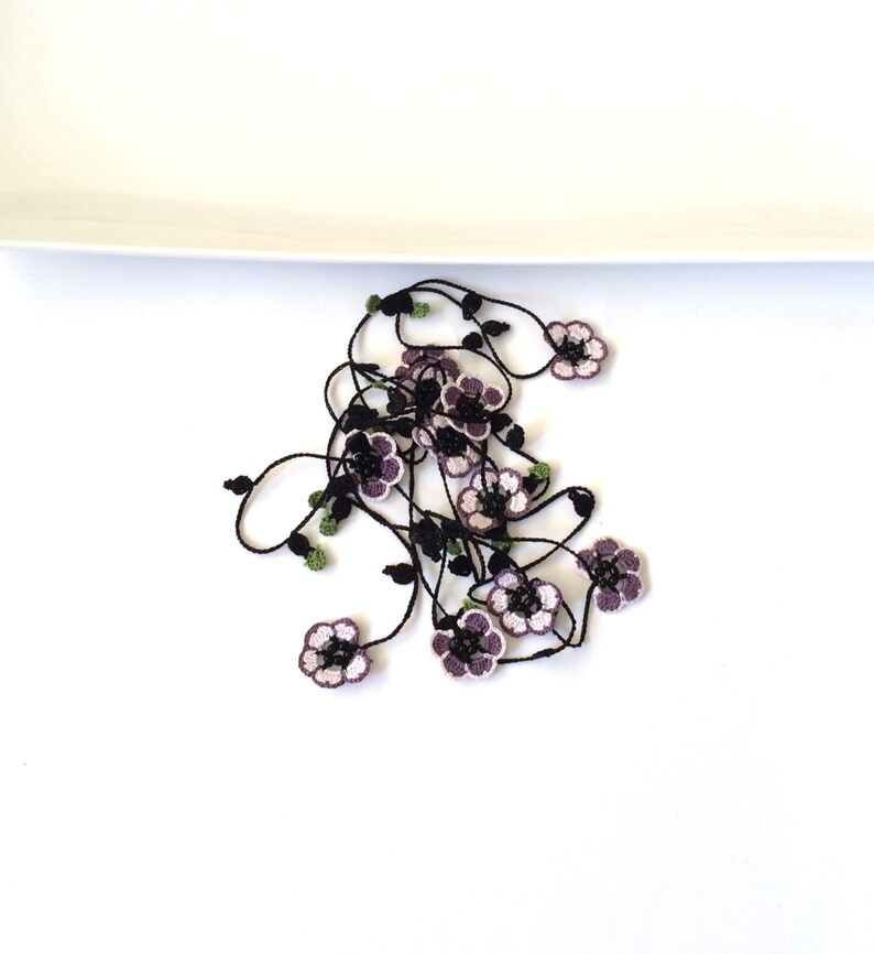 May include: A black crocheted necklace with purple and white flowers. The necklace is on a white surface.