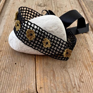 May include: A black crocheted headband with gold-coloured flower accents. The headband is made of a net-like material and features a black satin ribbon. The headband is resting on a white stone, set against a wooden background.