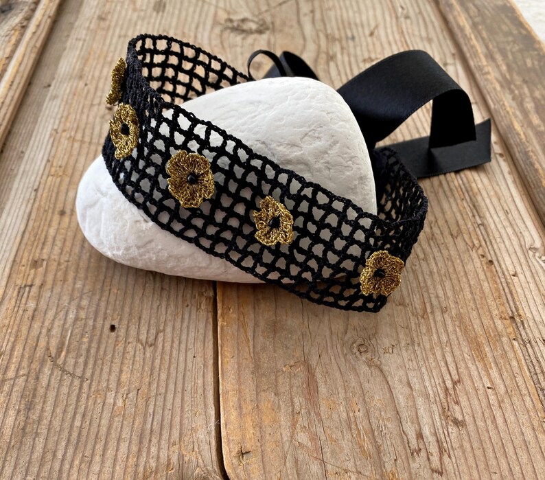 May include: A black crocheted headband with gold-coloured flower accents. The headband is made of a net-like material and features a black satin ribbon. The headband is resting on a white stone, set against a wooden background.