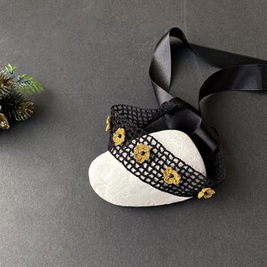 May include: A black crocheted headband with gold-coloured flower accents is draped over a white stone. A black satin ribbon is attached to the headband. The background is a dark grey surface.