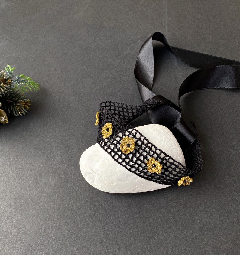 May include: A black crocheted headband with gold-coloured flower accents is draped over a white stone. A black satin ribbon is attached to the headband. The background is a dark grey surface.