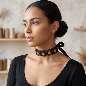 May include: A black choker necklace with a floral design. The choker is made of a mesh material and features gold-coloured flower accents. A black ribbon ties the necklace at the back. The person is wearing a black long-sleeve shirt.